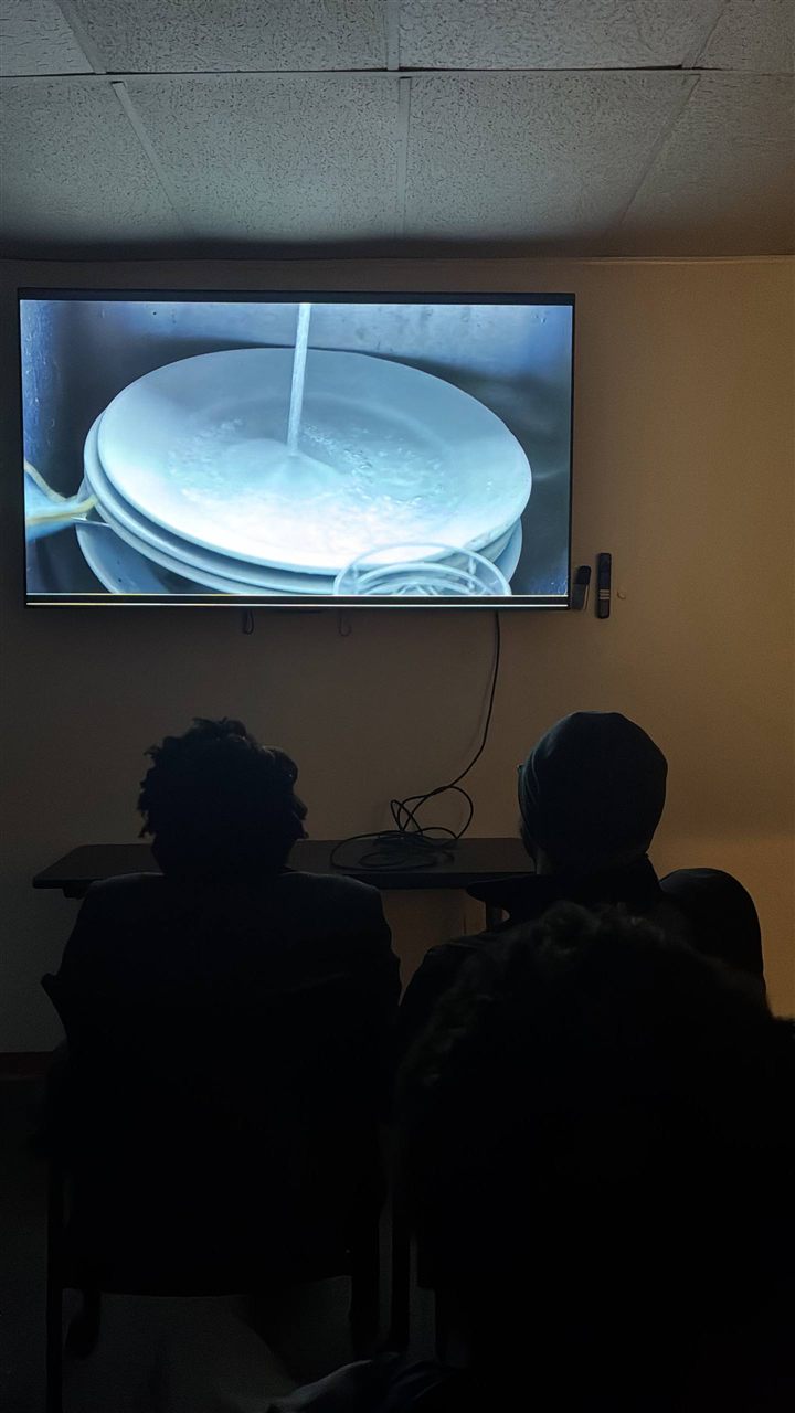 Two viewers from behind, a short film playing on the wall-mounted screen.