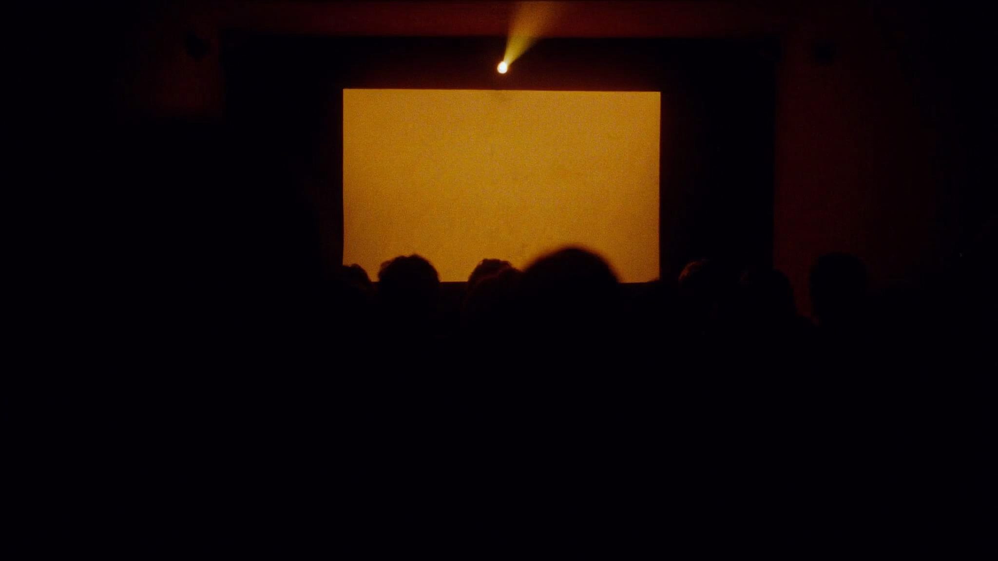 An intimate small theater room seen from the back, a single glowing screen of warm amber light at the far end, audience silhouettes from behind in the foreground rows.