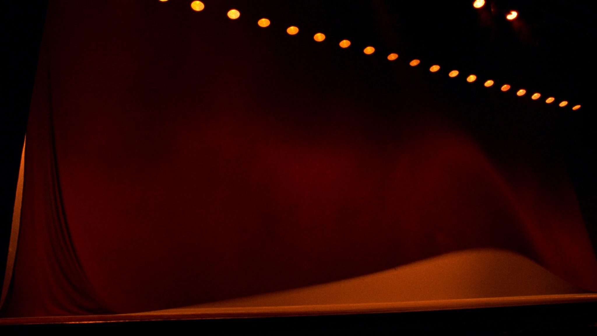 A heavy red velvet curtain draped against deep darkness, a row of warm theater bulbs glowing along the top edge.